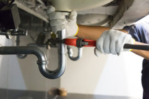 A plumber uses a wrench to loosen a silver pipe under a sink.