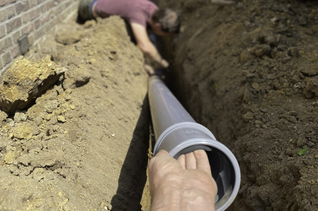 A man carries a sewer pipe and places it in the dirt foundation laid out for it.