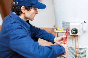 A plumber in a blue uniform uses a wrench to work on a water heater.