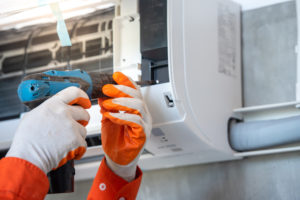 A man's orange gloves can be seen using a screwdriver to help repair an AC unit.