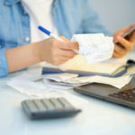 A woman sits at a desk with a calculator, laptop and pile of receipts. She is trying to factor in a bill's cost.